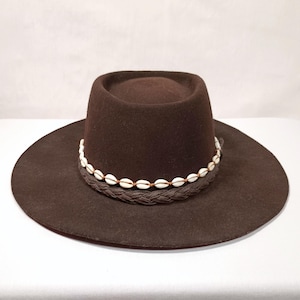 Op de afbeelding: Een bruine wollen fedora hoed met een gevlochten band en witte schelp accenten.