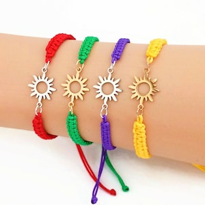 May include: Four woven cord bracelets in red, green, purple, and yellow. Each bracelet features a central metallic sun-shaped charm with radiating points. The cords are braided and adjustable, with loose ends hanging down from the main band.