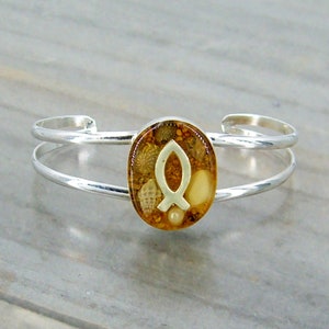 May include: Silver-tone cuff bracelet with an oval centerpiece. The centerpiece is filled with small seashells and sand, and features a silver fish symbol. The bracelet has a double-band design.