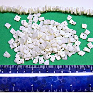 May include: A large pile of white, square-shaped shell beads with a hole in the center. The beads are scattered on a green surface with a ruler in the foreground.