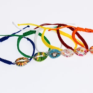 May include: Seven colorful braided bracelets with a shell charm. The charms are decorated with floral patterns in various colors. The bracelets are made of different colors of cord, including blue, green, white, yellow, orange, and red.