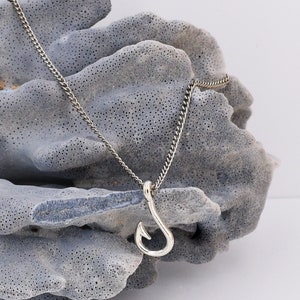 May include: A silver chain necklace with a silver hook pendant. The necklace is displayed on a gray textured surface.