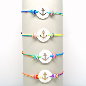 May include: Four colorful bracelets with white shell charms featuring gold anchor designs. The bracelets are layered on a white display stand.