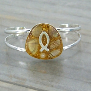 May include: Silver cuff bracelet with a clear resin charm featuring a silver fish symbol and embedded seashells. The charm is a light brown color.