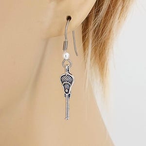 May include: Silver lacrosse stick charm earrings with a white pearl accent. The earrings are on a silver hook.