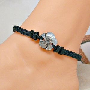 May include: A black braided ankle bracelet with a silver flower charm. The charm has a detailed design and is centered on the bracelet.