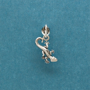 May include: A silver charm in the shape of a gecko lizard. The charm has a loop at the top for attaching to a bracelet or necklace.