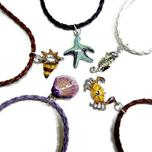 Op de afbeelding: Vijf verschillende lederen koordkettingen met verschillende emaille-charms, waaronder een schelp, een zeester, een krab, een zeepaardje en een schelp. De charms zijn allemaal zilverkleurig en hebben kleurrijke emaille-details.