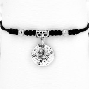 May include: A black cord bracelet with a silver charm in the shape of a sand dollar. The sand dollar charm has a star design in the center.