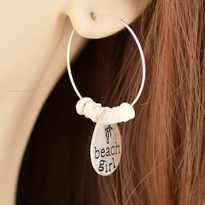 Peut inclure: Boucles d'oreilles créoles argentées avec des perles blanches et un pendentif en forme de goutte d'eau. Le pendentif porte les mots "beach girl" et un motif de palmier. Les boucles d'oreilles sont portées à l'oreille.