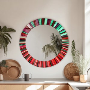 May include: A round mirror with a colorful, striped frame in shades of red, pink, and green. The mirror is hanging on a white wall in a kitchen setting.