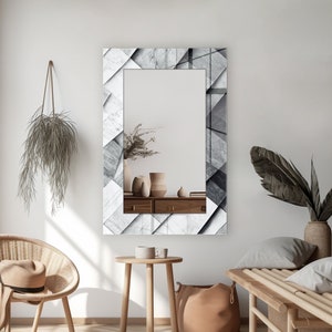 May include: A rectangular mirror with a white and gray geometric pattern frame. The mirror is hanging on a white wall and reflects a wooden dresser with vases and plants.