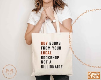 May include: A cream-colored canvas tote bag featuring the text BUY BOOKS FROM YOUR LOCAL BOOKSHOP NOT A BILLIONAIRE printed in a combination of orange and black block lettering. The bag has two shoulder straps and a rectangular shape.
