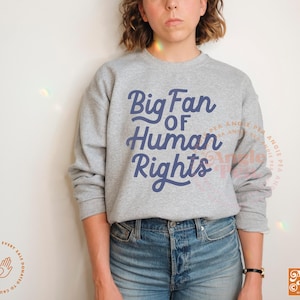 May include: A heather gray sweatshirt with the text "Big Fan of Human Rights" in navy blue script. The sweatshirt is paired with blue jeans. The design includes the Angie Pea logo.