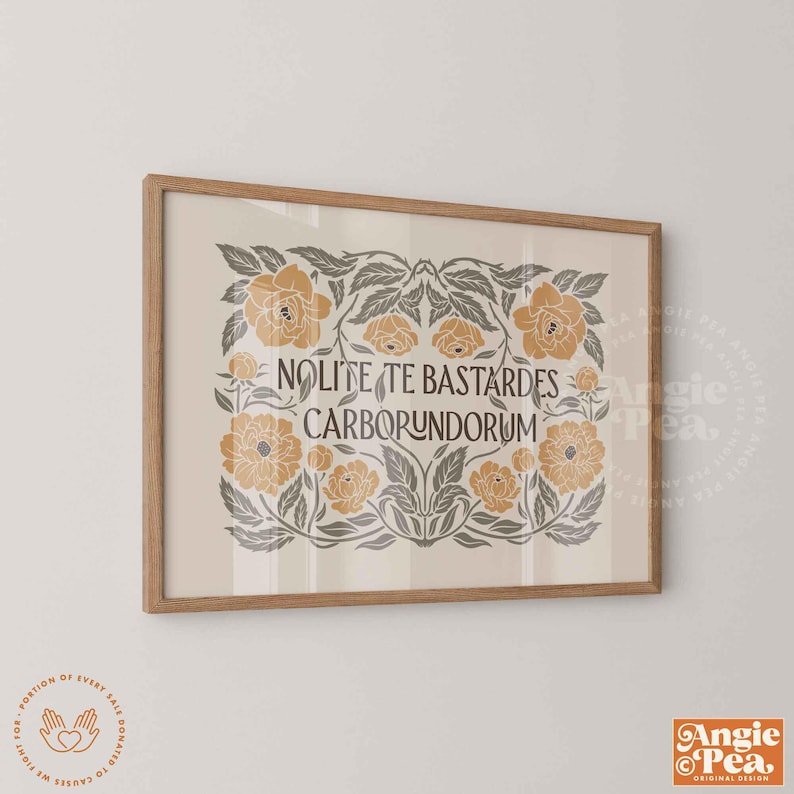 May include: A framed print with a light beige background and a floral design in shades of gold and gray. The text "Nolite Te Bastardes Carborundorum" is centered. The frame is a light brown color.
