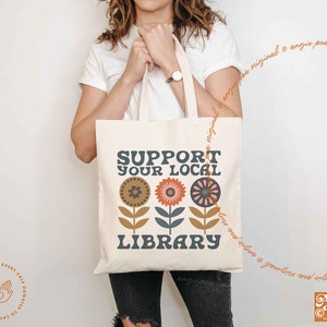 May include: A white tote bag with the text "Support Your Local Library" in blue and brown lettering. The bag features a design of three stylized flowers with orange, brown, and pink petals.