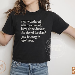 May include: A black cropped t-shirt with white text that reads "ever wondered what you would have done during the rise of fascism? you're doing it right now."