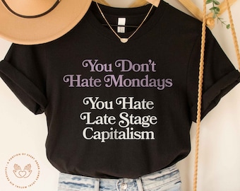 May include: Black t-shirt with the text "You Don't Hate Mondays You Hate Late Stage Capitalism" in white and purple lettering.