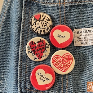 May include: A collection of round buttons with various designs and text, including "Love Over Hate," "igaf," and "Abolish Ice." The buttons are red, white, and black, and are pinned to a denim jacket.