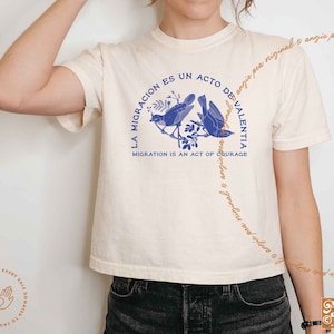 May include: A white cropped t-shirt with a blue bird graphic and the text "La Migracion es un acto de Valentia" in Spanish and "Migration is an act of courage" in English.