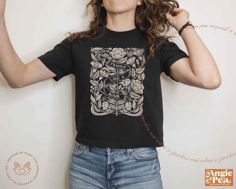 K&ouml;nnte beinhalten: Ein schwarzes, bauchfreies T-Shirt mit einem floralen Design und dem Text "Fragile Like A Bomb".