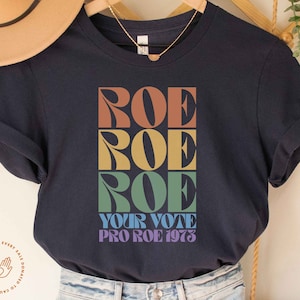 May include: A navy blue t-shirt with a graphic design of the word "ROE" repeated three times in a rainbow color scheme. The text "YOUR VOTE PRO ROE 1973" is printed below the word "ROE".