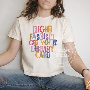 May include: A white t-shirt with a colorful graphic that reads "Fight Fascism Get Your Library Card".