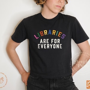 以下が含まれることがあります： 黒いクルーネックTシャツ。テキスト「LIBRARIES ARE FOR EVERYONE」が虹色でプリントされています。半袖Tシャツです。