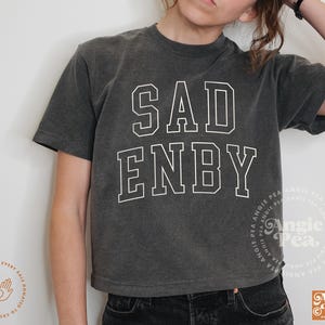 May include: A dark gray cropped t-shirt with the text "SAD ENBY" printed in white on the front.