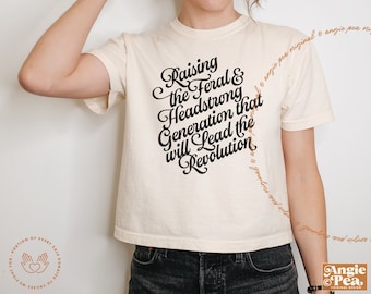 以下が含まれることがあります： 黒い文字で「Raising the Feral & Headstrong Generation that will Lead the Revolution」と書かれた白いクロップドTシャツです。