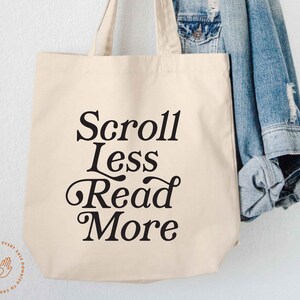May include: A cream-colored canvas tote bag featuring the black printed text Scroll Less Read More in a stylized serif font. The bag has two top handles and a simple rectangular shape, presented with a blue denim jacket hanging nearby.