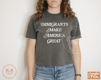 Op de afbeelding: Een donkergrijs T-shirt met de tekst "Immigrants Make America Great" in wit gedrukt. Het shirt is een cropped style en wordt door iemand gedragen.