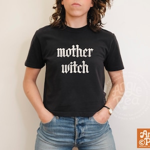 以下が含まれることがあります： 胸元に白いゴシック体のフォントで「mother witch」というテキストがプリントされた、黒の半袖Tシャツ。クルーネックのデザインで、ブルーのデニムジーンズと組み合わされており、シンプルでカジュアルな外観を呈しています。