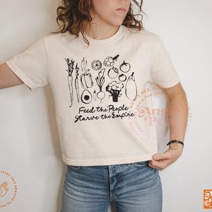 May include: Cream-colored t-shirt with a black graphic of various vegetables and the text "Feed the People, Starve the Empire." The shirt is a crew neck and is paired with blue jeans. The shirt has short sleeves.