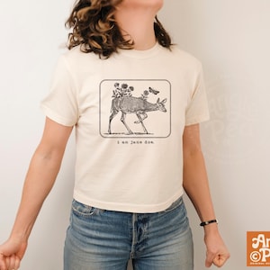 Op de afbeelding: Een crèmekleurig T-shirt met korte mouwen en een zwarte lijntekening van een hert met bloemen op de rug en een vlinder. Het ontwerp staat in een afgeronde rechthoek met de tekst "i am jane doe." eronder gedrukt.