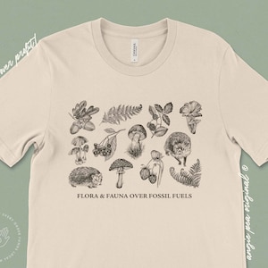May include: A beige t-shirt with a black and white illustration of various flora and fauna, including mushrooms, berries, ferns, and hedgehogs. The text "FLORA & FAUNA OVER FOSSIL FUELS" is printed below the illustration.