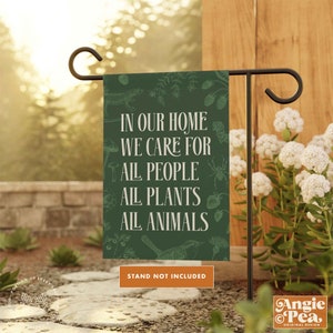 May include: Green garden flag with white text that reads "In our home we care for all people all plants all animals". The flag has a green background with a variety of small, white, and red illustrations of plants and animals. The text "Stand not included" is printed in white on a brown rectangle at the bottom of the flag.