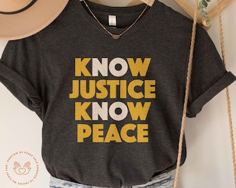 May include: A dark gray t-shirt with the words "KNOW JUSTICE KNOW PEACE" printed in yellow and white letters. The shirt is on a hanger and has a portion of a brown hat and a rope hanging in the background.