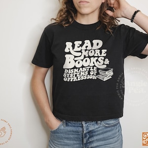 May include: A black t-shirt with white text that reads "Read More Books & Dismantle Systems of Oppression". The text is in a retro font style. There are three stacked books to the right of the text.