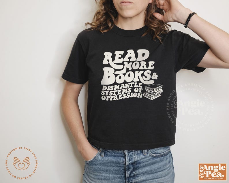 May include: A black t-shirt with white text that reads "Read More Books & Dismantle Systems of Oppression". The text is in a retro font style. There are three stacked books to the right of the text.