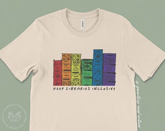 Könnte beinhalten: Ein beiges T-Shirt mit einer regenbogenfarbenen Grafik von gestapelten Büchern. Die Grafik lautet "Keep Libraries Inclusive".