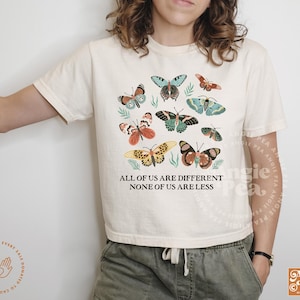 May include: A white t-shirt with a colorful butterfly design and the text "All of us are different, none of us are less".