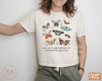 Può includere: Una maglietta bianca con un design di farfalla colorato e il testo "All of us are different, none of us are less".