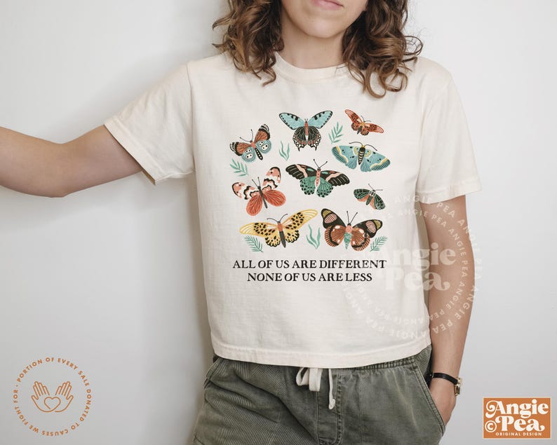May include: A white t-shirt with a colorful butterfly design and the text "All of us are different, none of us are less".