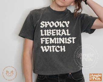 May include: Dark gray t-shirt with the words "SPOOKY LIBERAL FEMINIST WITCH" in white gothic lettering. The shirt is a cropped style and has a vintage, worn look. The shirt is part of the Angie Pea original design collection.