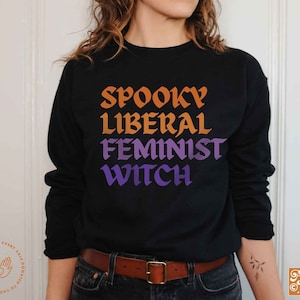 May include: Black sweatshirt with a graphic print that reads "Spooky Liberal Feminist Witch" in orange, yellow, purple, and blue lettering.