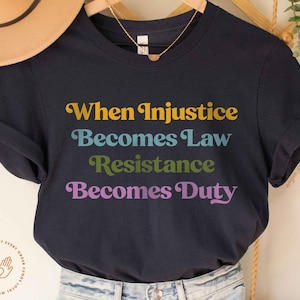 以下が含まれることがあります： カラフルなブロック体で「When Injustice Becomes Law Resistance Becomes Duty」と書かれたネイビーブルーのTシャツです。