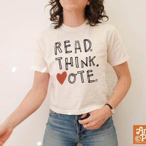 May include: A cream-colored short-sleeve t-shirt featuring black hand-drawn style lettering that reads READ. THINK. VOTE. with a solid red heart icon replacing the letter V in the word VOTE. The shirt has a crew neckline and a standard fit.