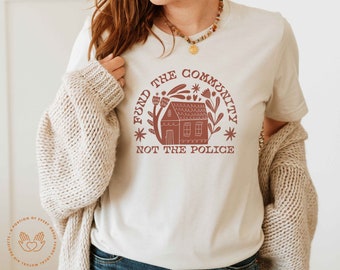 May include: A white t-shirt with a red graphic design that says "Fund the Community, Not the Police". The graphic features a house with a red roof and green trees and flowers.