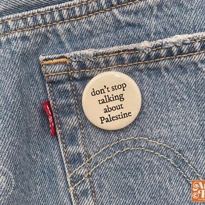 May include: A light blue denim jean pocket with a round, off-white button pinned to it. The button has the text "don't stop talking about Palestine" in black. The Levi's red tag is visible.